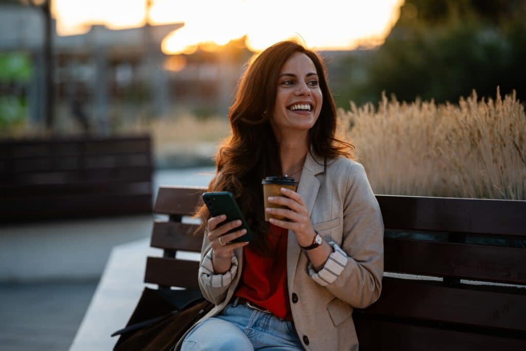 Woman,Sitting,On,A,Park,Bench,,Holding,A,Smartphone,And